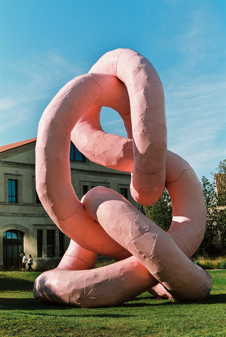 Arles, sculpture tubes roses entrelacés, pelouse verte, ciel bleu légèrement nuageux, contraste doux